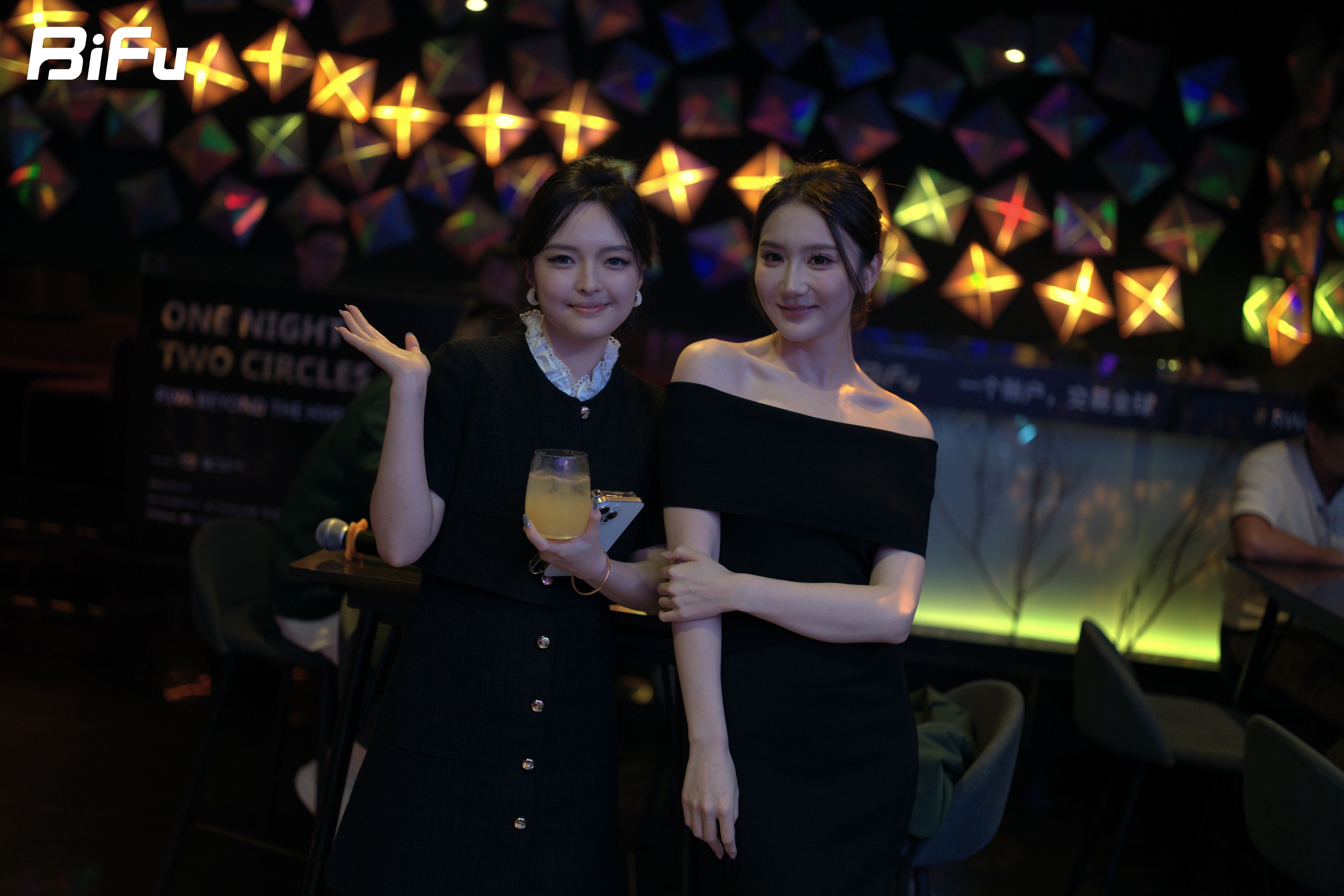 The picture shows two women standing together at the BiFu Token2049 RWA theme dinner and wine party. The woman on the left is wearing a black dress with a white collar and holding a drink, while the woman on the right is in a black off-the-shoulder dress. They are smiling and posing for the camera. The background features a colorful geometric pattern wall with the text "ONE NIGHT TWO CIRCLES" and "BiFu" displayed. This photo reflects the social interaction and atmosphere of the event where attendees deeply discussed, exchanged views, and connected.