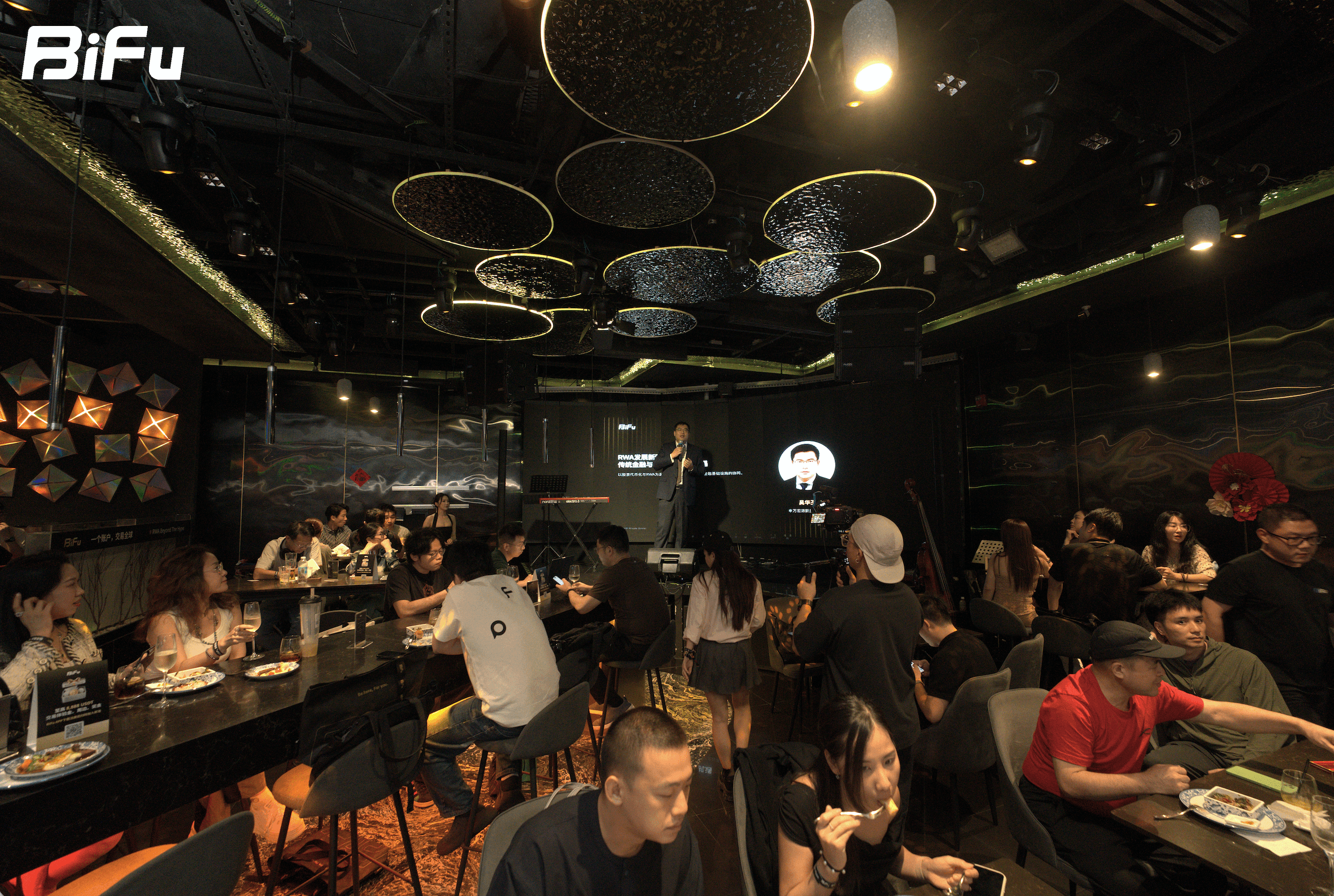 The picture shows the scene of BiFu's first BiFu Token 2049 RWA themed dinner and wine party. There are many people in the room, some sitting at tables eating and drinking, and some standing. On the stage, there is a person speaking. The ceiling has some circular lights. The background wall has some decorations.