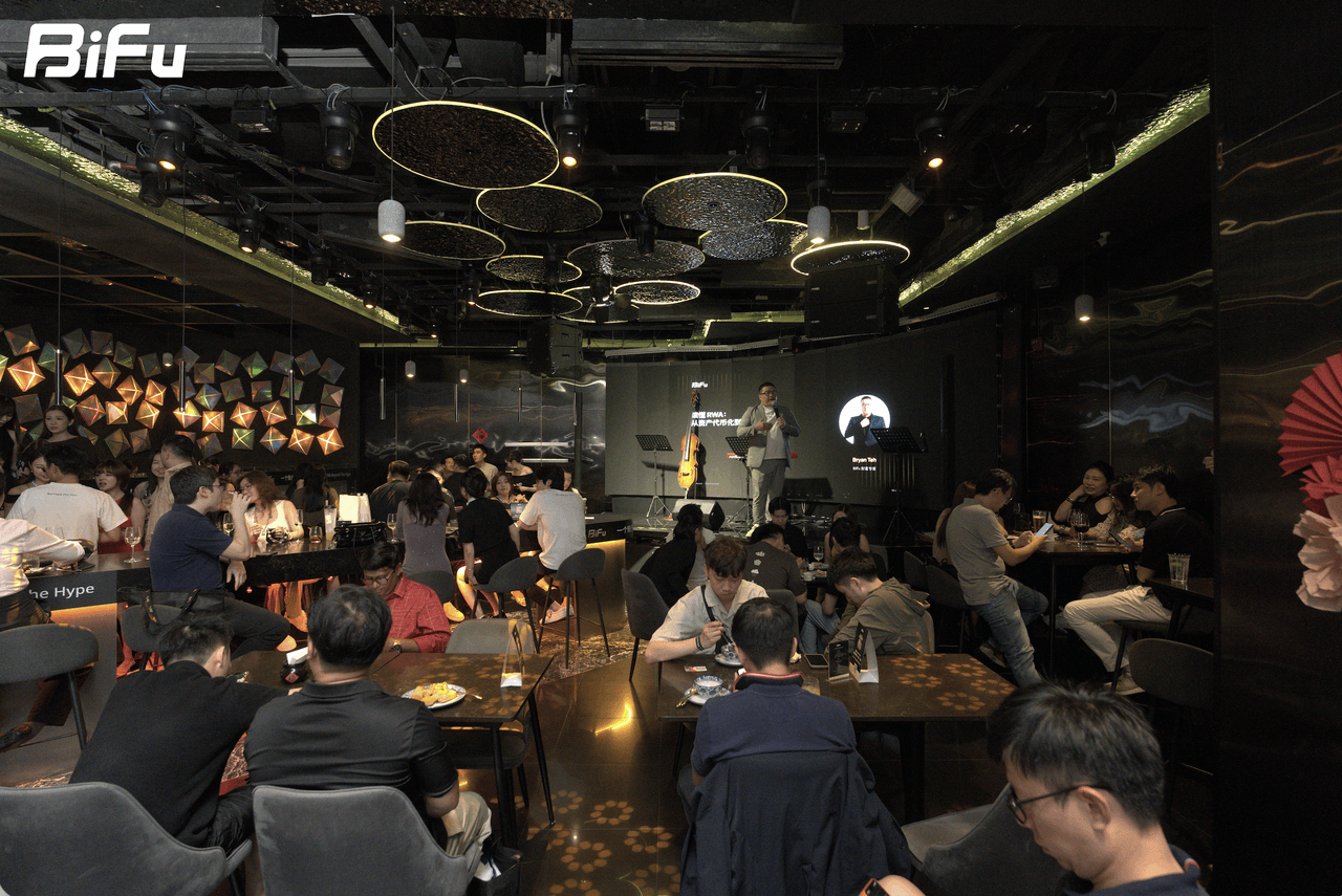 The picture shows the scene of BiFu's RWA-themed dinner and reception at Token2049. There are many people sitting at tables in a dimly lit venue with a modern ceiling design. A person is standing on a stage with a microphone, possibly giving a speech. The background screen displays some content, and there are colorful decorations on the left side of the stage. This corresponds to the context that the event was held with a focus on RWA, and the speaker pointed out issues like fragmented user accounts and the need for globalized opportunities.