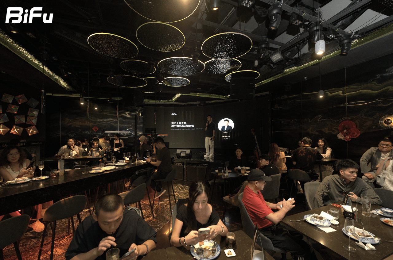 The picture shows the scene of BiFu's RWA-themed dinner and reception at Token2049. There are many people sitting at tables in the venue, some eating and some looking at their phones. A person is standing on the stage in front of a screen, and the screen displays some text. The venue has a dark ceiling with some lights and decorations.