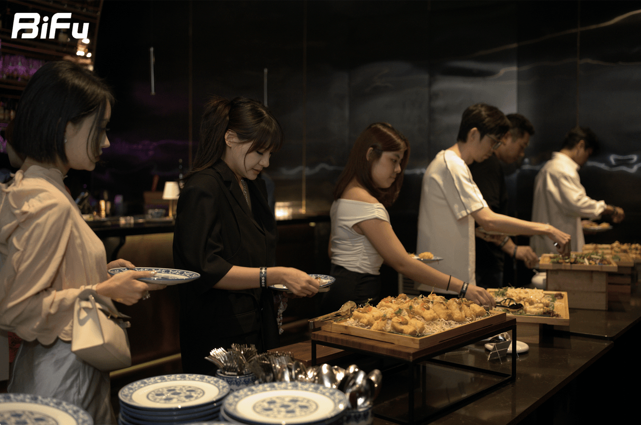 The picture shows a scene from the BiFu RWA theme dinner and wine party. Several people are standing at a buffet table, serving themselves food. There are various dishes on the table, including some with blue patterns. The background has some dim lighting and a black wall. This image corresponds to the context which mentions the BiFu RWA theme dinner and wine party, where four speakers discussed "What is missing for RWA to truly land" from four perspectives.