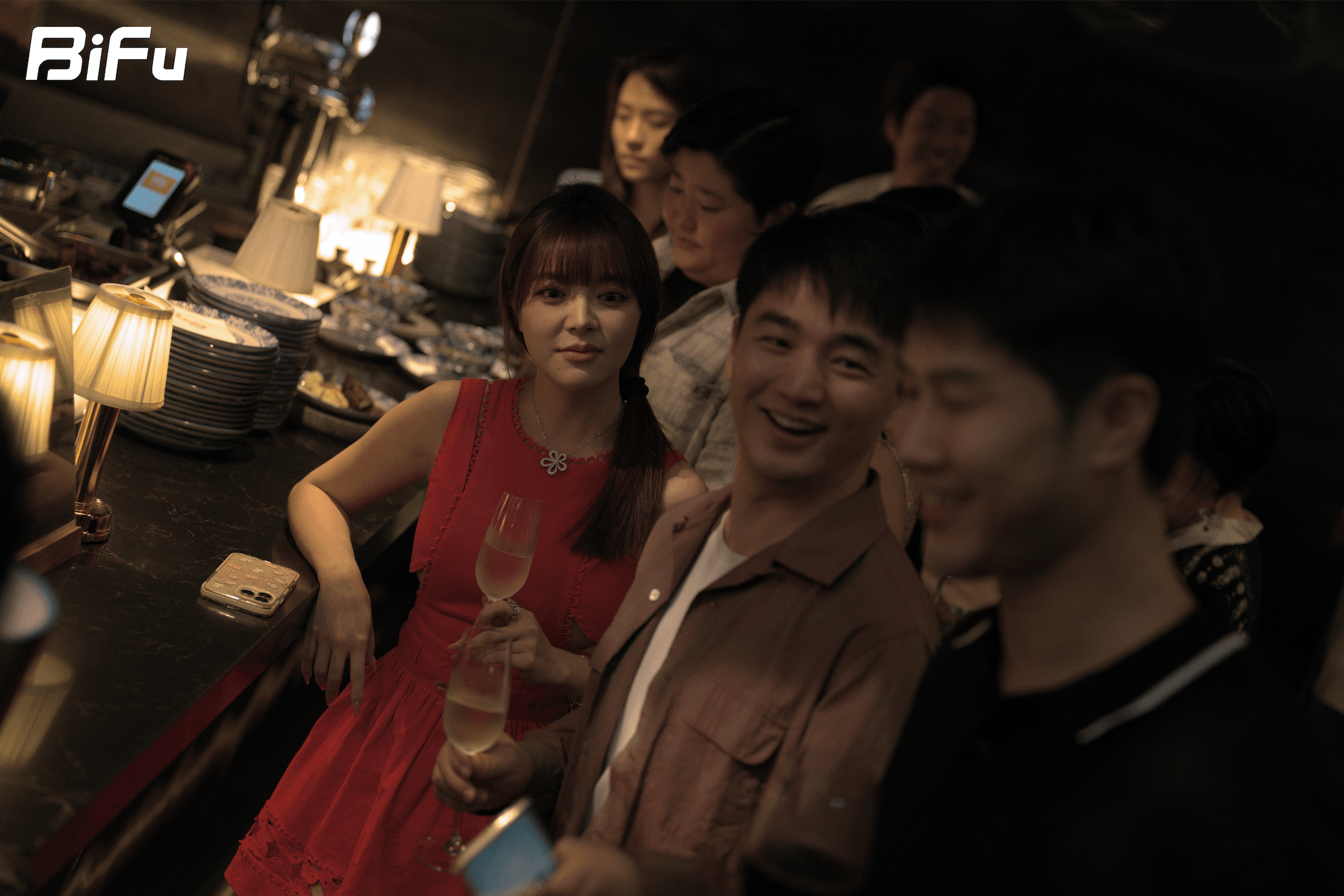 The picture shows a scene from BiFu's Token2049 RWA theme dinner and wine party. There are several people in the photo, including a woman in a red dress holding a glass of wine and a man in a brown shirt smiling. The background has some lamps and other items, and the BiFu logo is in the upper left corner. This photo corresponds to the context which mentions the dinner adopted a定向邀请制 with attendees from various parties related to RWA, and the closed and small-scale setting to facilitate industry exchanges.