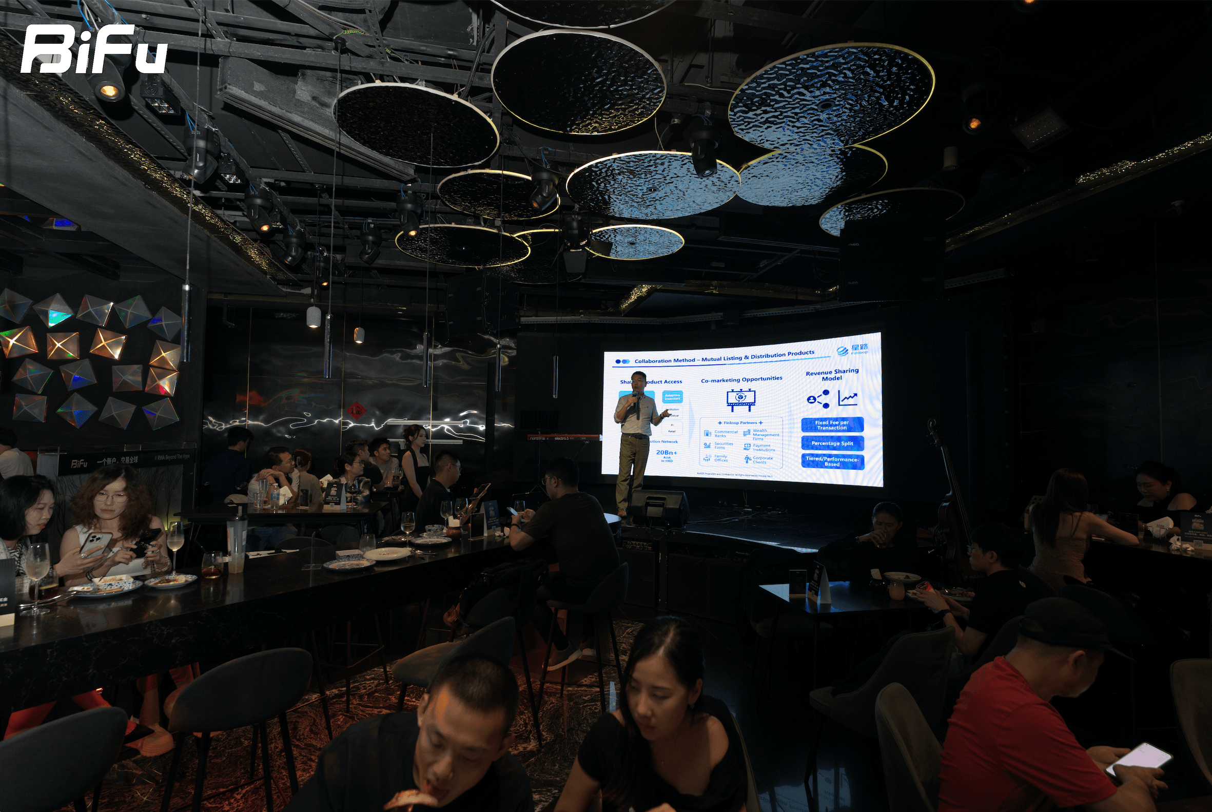 The picture shows the scene of BiFu's first show at Token2049 RWA themed dinner and wine party. There are many people sitting at tables in the venue, and a speaker is standing in front of a large screen. The screen displays some content related to RWA business. The venue has a dark ceiling with some lights, and there are some decorations on the wall.