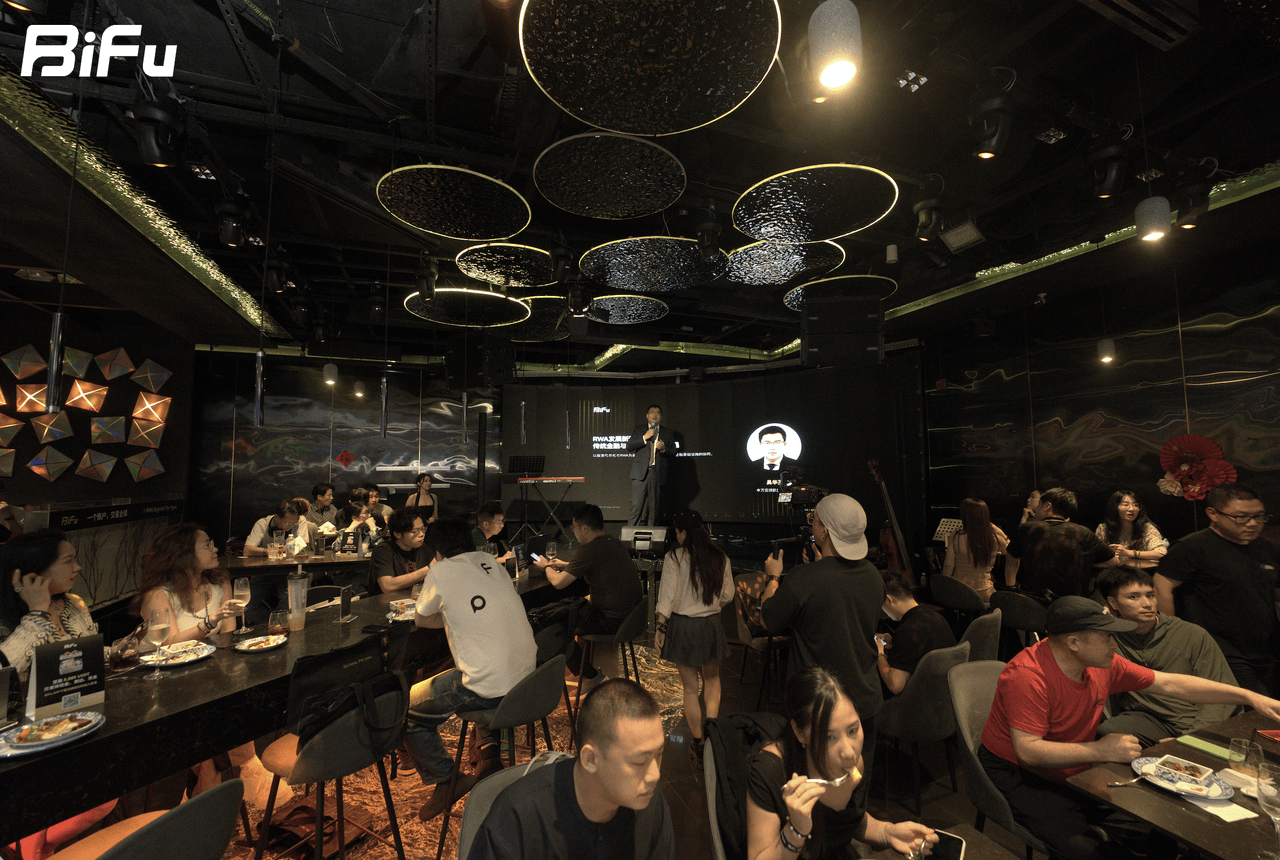 The image shows the scene of BiFu's Token2049 RWA theme dinner and reception. There are many people sitting at tables in a dimly lit room with a black ceiling and decorative lights. A person is standing on a stage with a screen behind them, and there are some people standing and others sitting around the tables. The BiFu logo is displayed in the top left corner of the image.
