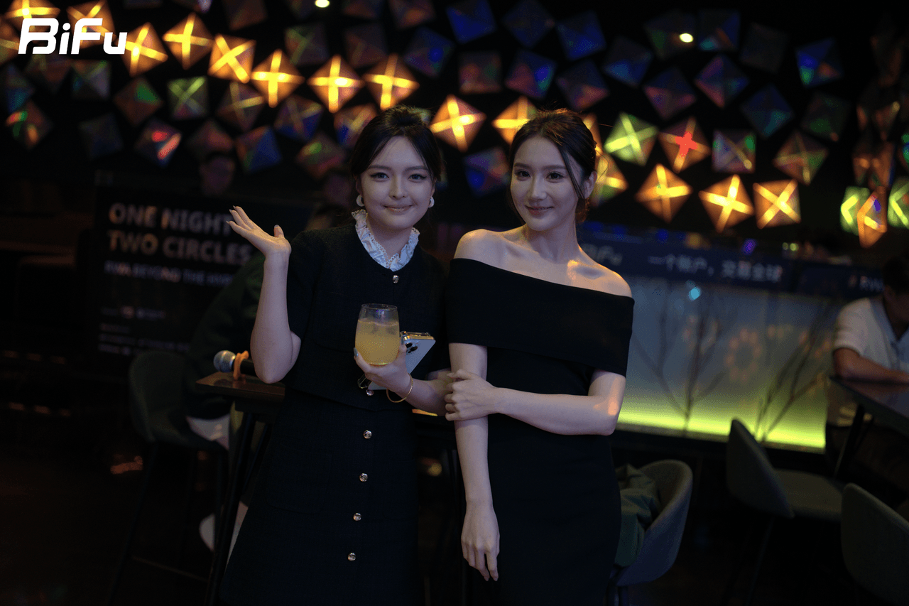 The picture shows two women standing together at the BiFu RWA theme dinner and reception of Token2049. They are both wearing black dresses, with one holding a drink in her hand. The background features a colorful, geometric-patterned wall with the text "ONE NIGHT TWO CIRCLES" and "BiFu" visible. This scene reflects the social interaction and networking atmosphere of the event where participants discussed projects, challenges, and future collaboration directions.