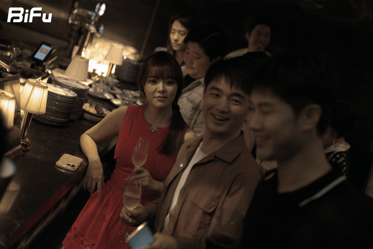The picture shows a scene from BiFu's Token2049 RWA-themed dinner and reception. There are several people gathered, with one woman in a red dress holding a glass of wine and smiling, and others around her engaged in conversation. The background features a bar area with lamps and some stacked items. This image corresponds to the context which mentions the event was held in a relaxed atmosphere where attendees discussed serious topics like the practical issues of on-chain asset ownership.