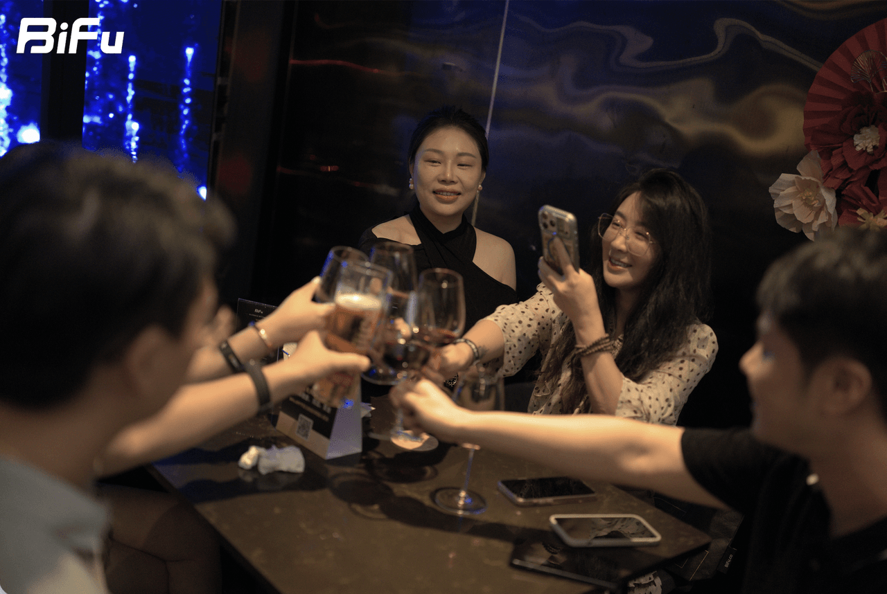 The picture shows a group of people toasting with wine glasses at a table during the BiFu RWA theme dinner and reception at Token2049. There are several wine glasses on the table, and one person is taking a photo with a phone. The background has a dark, decorative wall with some lights and a red flower decoration. The BiFu logo is visible in the top left corner of the image.