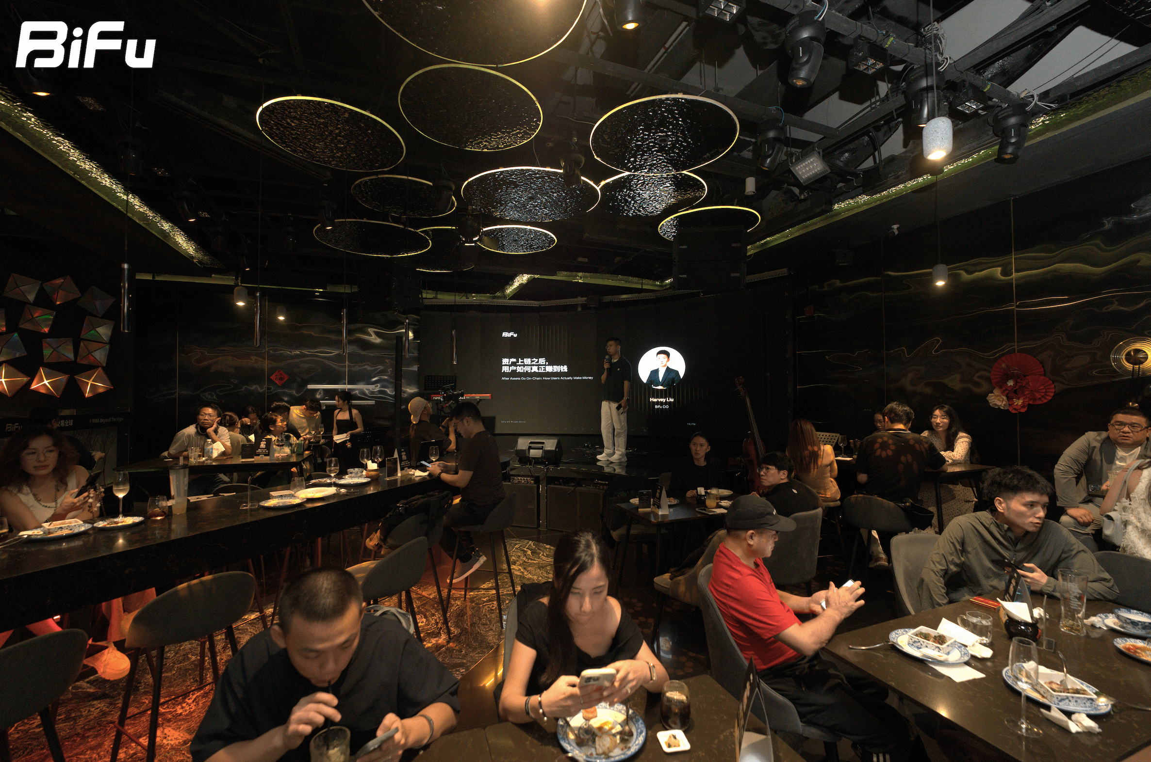 The picture shows the scene of BiFu's Token2049 RWA themed dinner and wine party. There are many people sitting at tables in a dimly lit room with a stage at the front. A person is standing on the stage, and there is a screen displaying some content. The ceiling has circular light fixtures, and there are some decorations around. This event is mentioned in the context where BiFu's CEO introduced the platform's boundaries, emphasizing that the platform does not guarantee profits and that the path to profits is determined by underlying assets and the market.