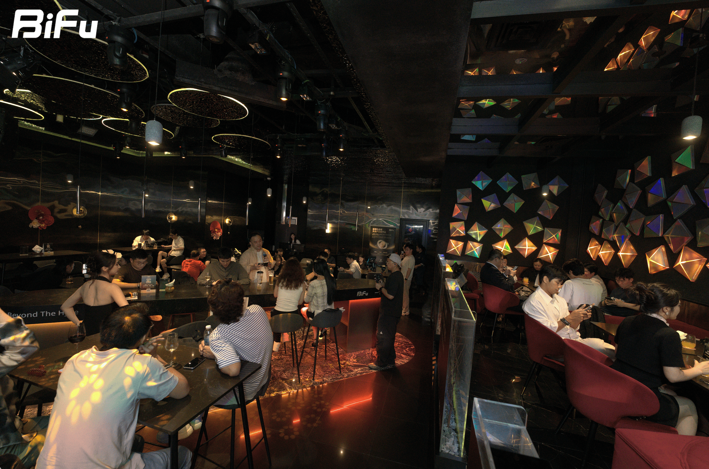 The picture shows the scene of BiFu's Token2049 RWA theme dinner and wine party. There are many people sitting at tables and chairs in the dimly lit venue, some are chatting, some are looking at their phones. The background has colorful geometric patterns on the wall and hanging lights. The BiFu logo is displayed in the upper left corner of the image. This event is mentioned in the context where BiFu held a show and the four speakers discussed the same problem from four angles.