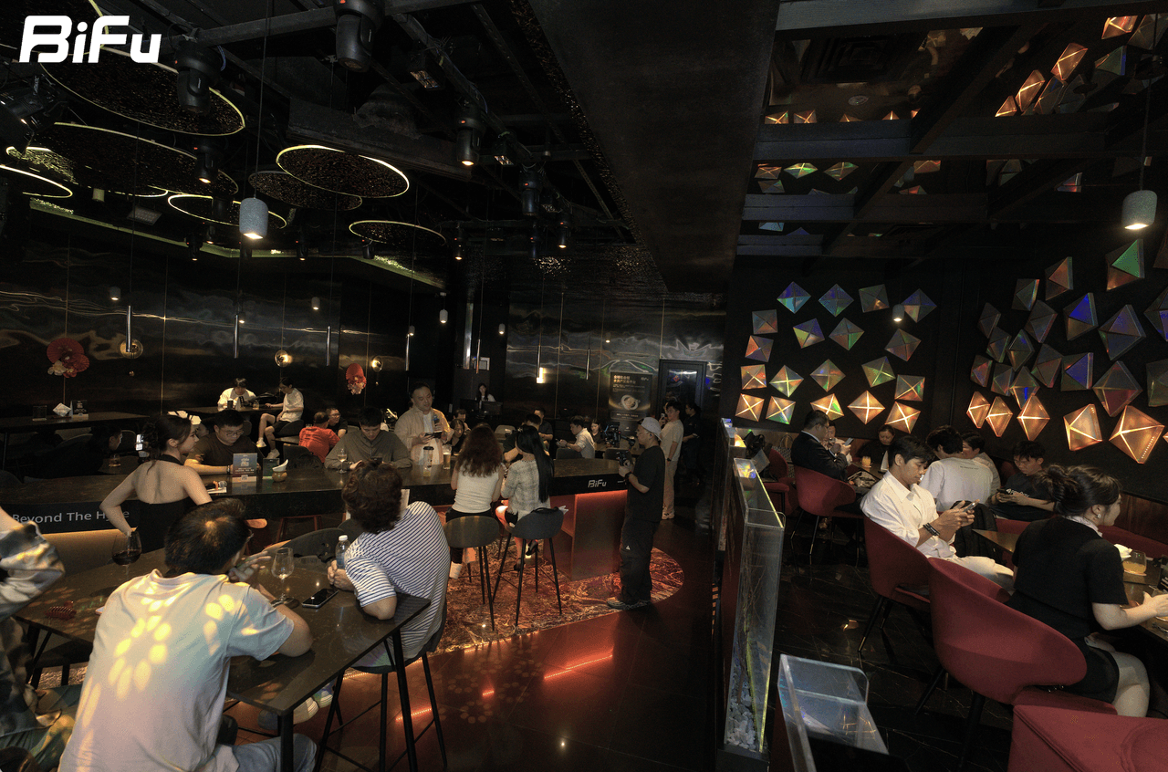 The picture shows the scene of BiFu's Token2049 RWA-themed dinner and reception. There are many people sitting at tables and standing in a dimly lit venue with a modern interior, including colorful geometric patterns on the walls and hanging lights. The BiFu logo is displayed in the top left corner of the image. This corresponds to the context which mentions that the event was held with 4 speakers discussing the same question from 4 perspectives.